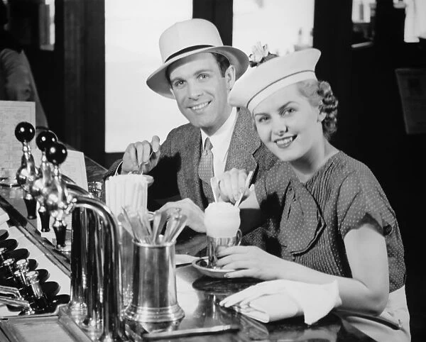 Man and woman in fancy hat drinking ice cream soda (B&W), portrait