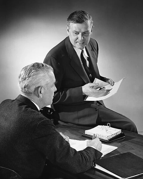 Two men talking, one sitting on desk
