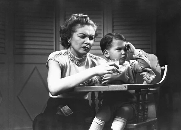 Mother giving glass of milk to son (2-3) sitting in high chair, (B&W)
