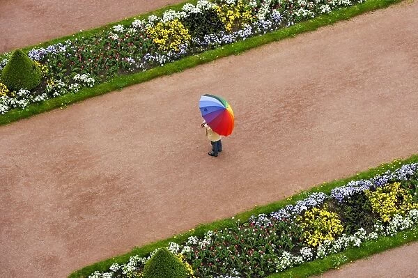 Person with umbrella in rainbow colors on a red gravel path between flower beds in the castle gardens, Fuldaer Stadtschloss City Palace, Fulda, Hesse, Germany