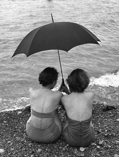 Rainy Holiday; Two women sheltering under their umbrella as they sit on the beach at Torquay in the rain