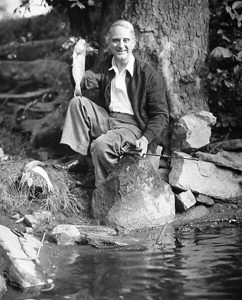 Senior man sitting by water, holding up fish (B&W), portrait