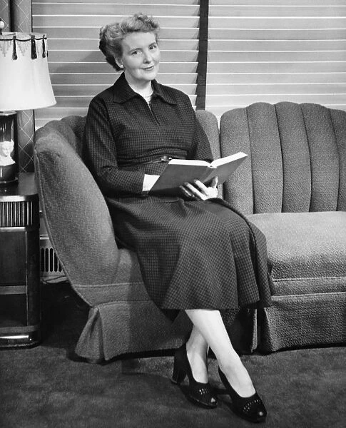 Senior woman sitting on sofa, holding book (B&W), portrait