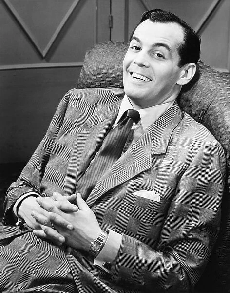 Smiling man in full suit reclining on chair, (B&W), portrait