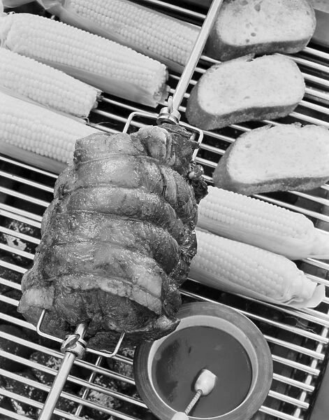 Spit roasting of ham on barbecue grill with corn cobs, close-up