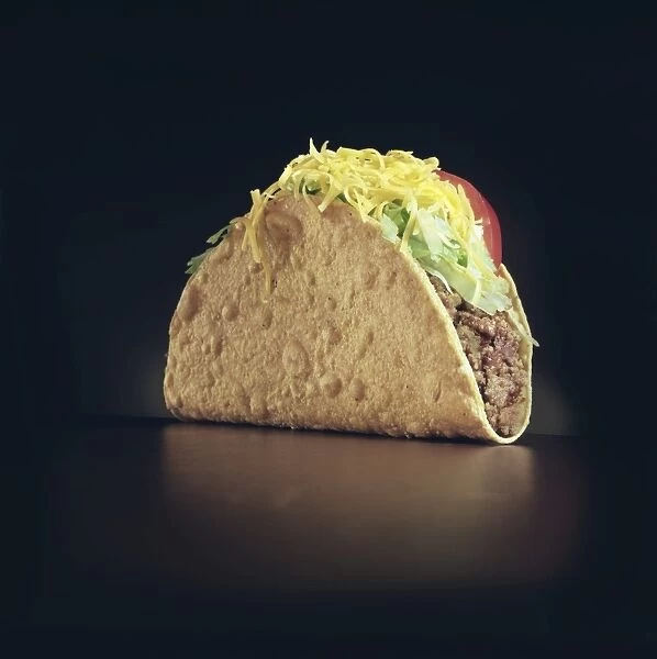A Taco. View of a meat taco with lettuce and tomatos on a black background, January 1989