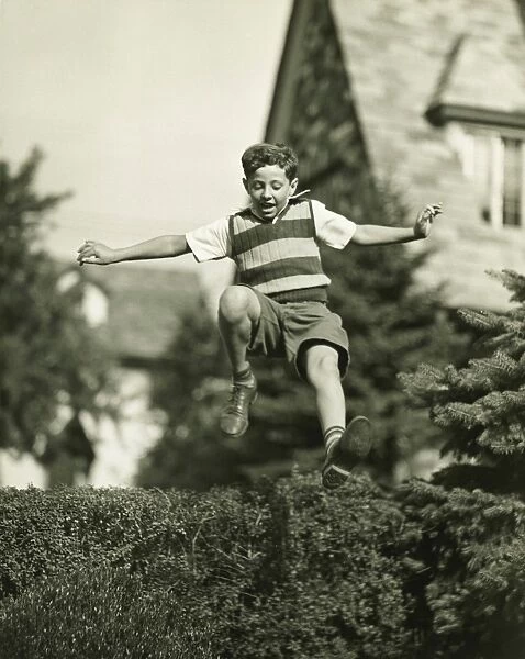 Teenage boy (13-14) leaping over hedge, (B&W), low angle view