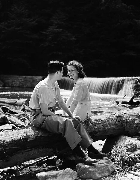 Teenage couple sitting on fallen log by side of creek, talking