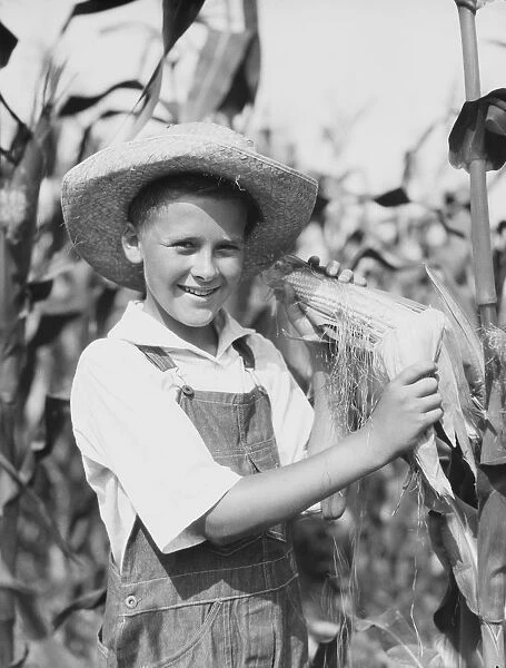 Teenage farm boy wearing bib overalls and straw hat, standing in corn field, holding corn cob