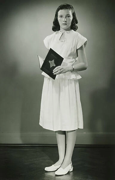 Teenage girl (13-14) in white dress standing in studio with open book, (B&W)