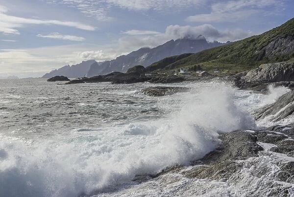 Waves breaking on the rocky coast, cottages and rugged mountains at the back, Flakstad, Lofoten, Nordland, Norway