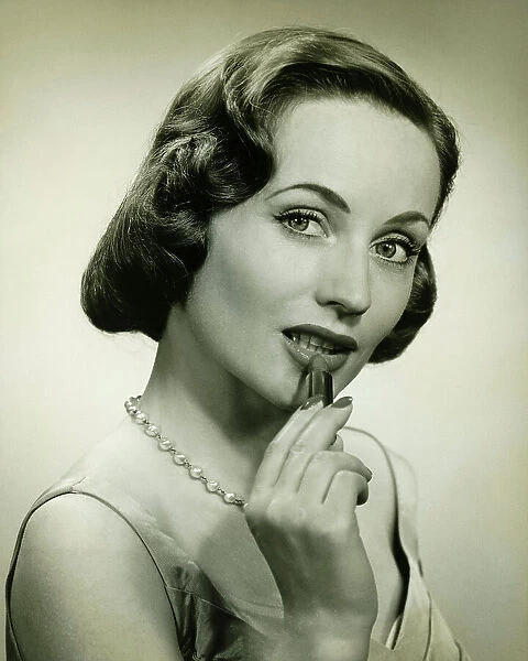 Woman applying lipstick in studio, (B&W), portrait