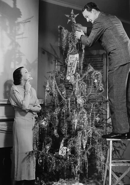 Woman assisting man placing star on top of Christmas tree, (B&W)