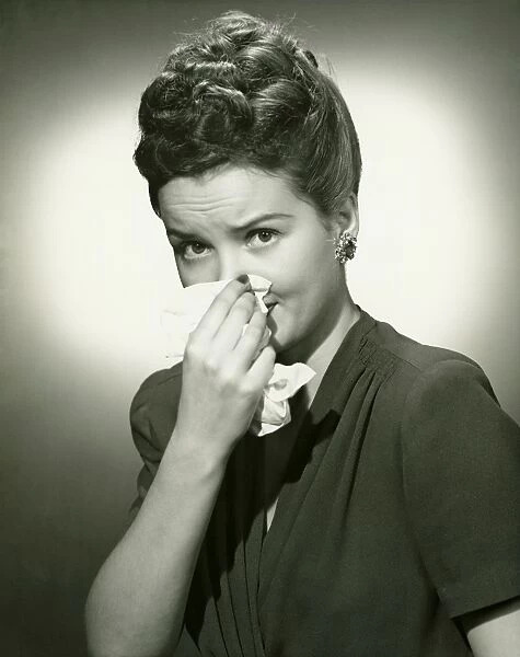 Woman blowing nose on tissue in studio, (B&W), portrait