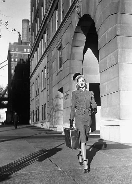 Woman carrying suitcase walking on sidewalk, (B&W)