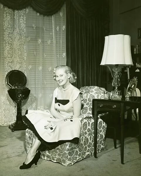 Woman in fashionable dress sitting on armchair in living room, (B&W), portrait