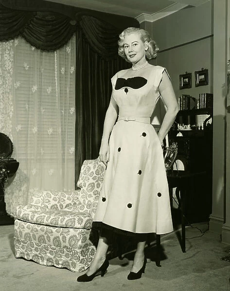 Woman in fashionable dress standing by armchair in living room, (B&W), portrait