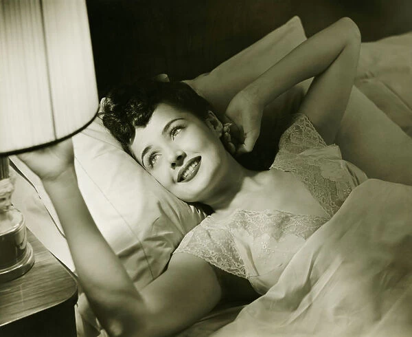 Woman lying on bed, turning off lamp on night table, (B&W), elevated view
