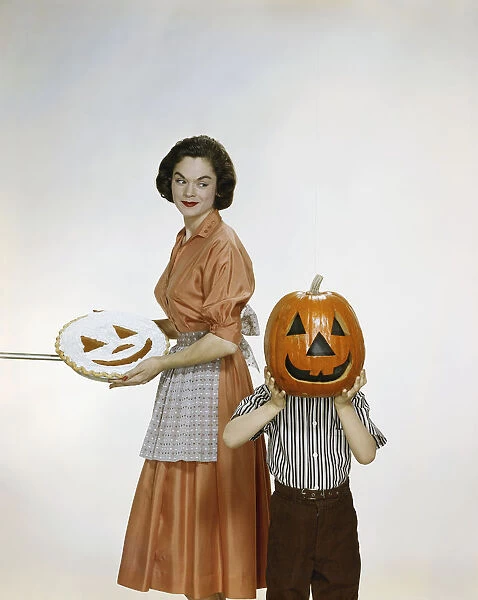 Woman with pie looking at child holding halloween pumpkin, smiling