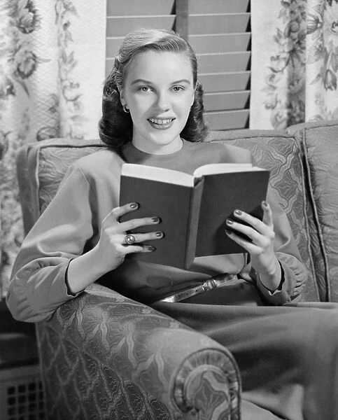 Woman reading book in living room, (B&W), portrait
