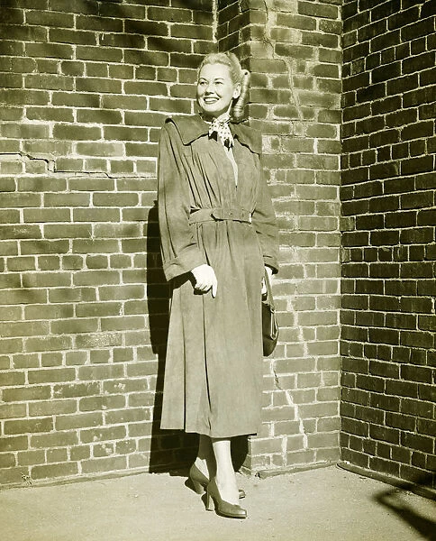 Woman wearing mackintosh standing at brick wall outdoors, (B&W)