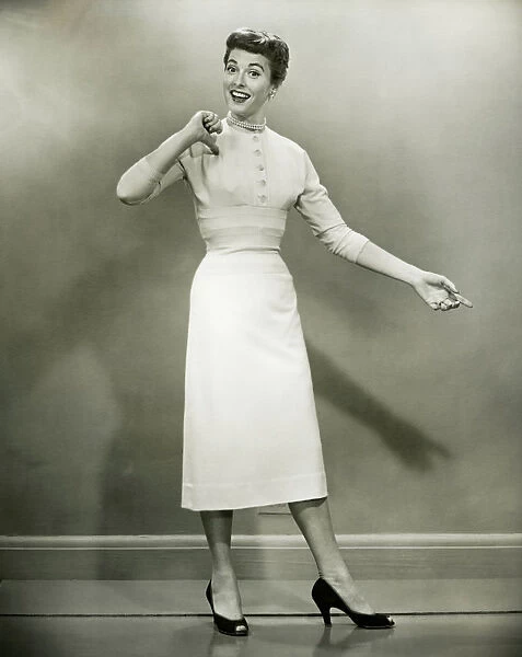 Woman in white dress pointing to herself in studio, (B&W), portrait