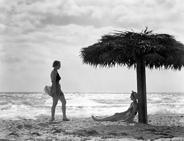 Two women under palm thatch umbrella on beach, Florida