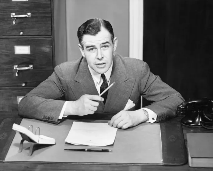 Framed Print : Young businessman sitting at small desk, holding pencil, (B&W)