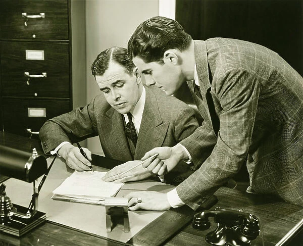 Two young businessmen talking at small desk, (B&W)