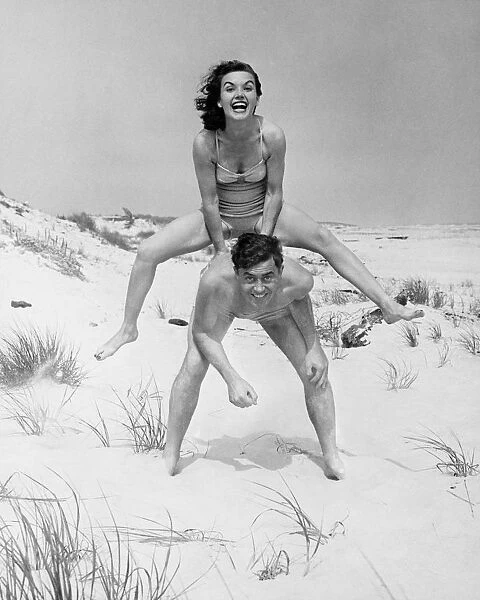 Young couple on beach, woman leap-frogging man, (B&W), portrait