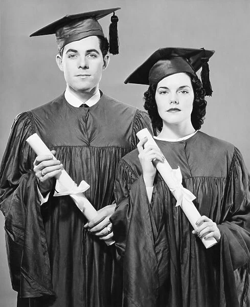 Young couple in graduate dress holding diplomas, posing in studio, (B&W), portrait