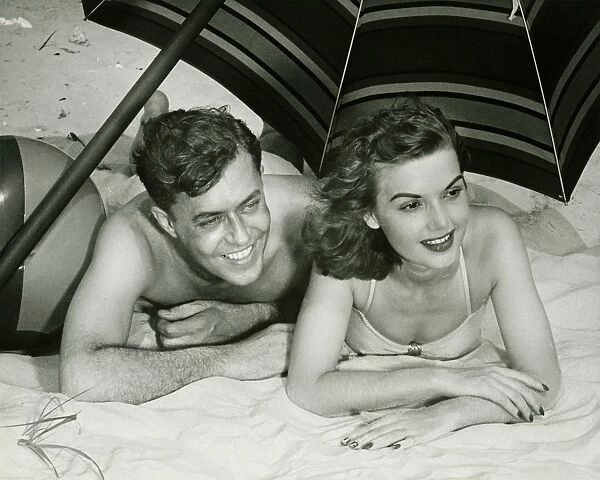 Young couple lying on sand under umbrella, smiling, (B&W)