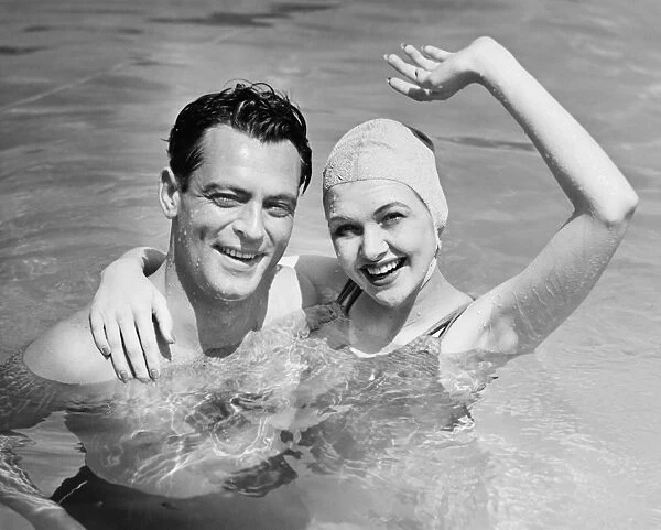 Young couple standing in pool, woman waving hand, (B&W), portrait