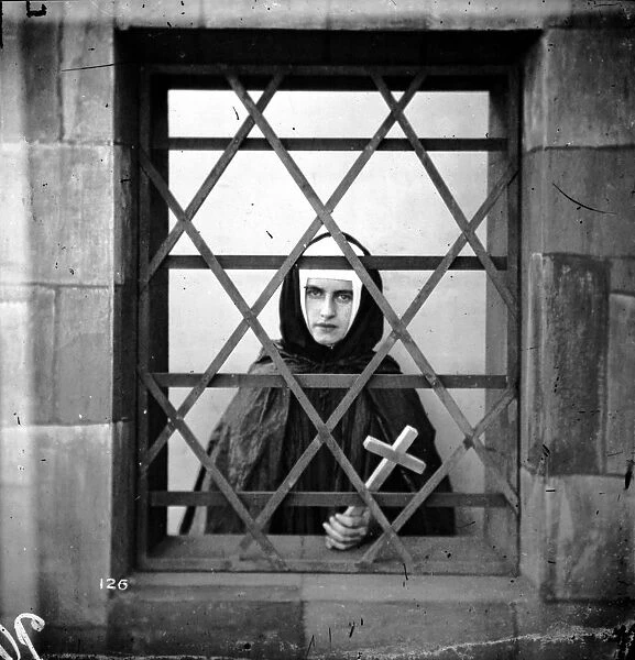 Young Nun. circa 1865: A young nun holding a cross whilst looking through a barred window