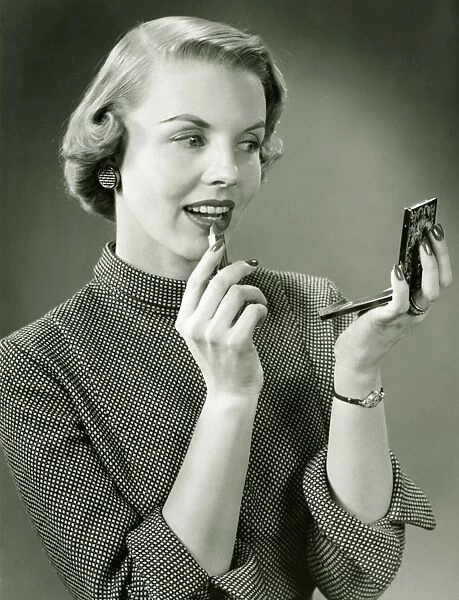 Young woman applying lipstick, looking at powder compact mirror, (B&W), close-up