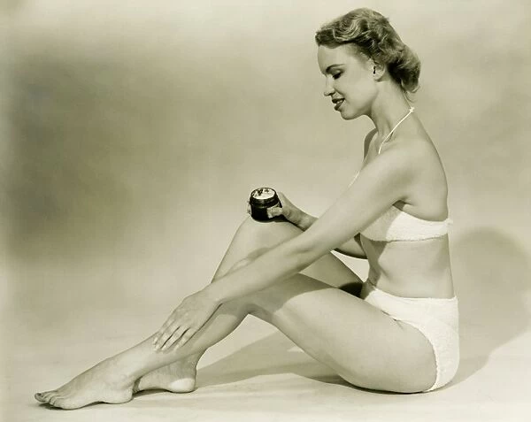 Young woman in bikini sitting, putting cream on leg, (B&W)