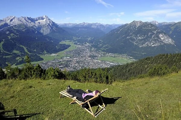Young woman in a deck chair, view of Garmisch-Partenkirchen, Zugspitze mountain, Jubilaeumsgrat ridge and Alpspitze mountain as seen from Wank mountain, Upper Bavaria, Bavaria, Germany, Europe