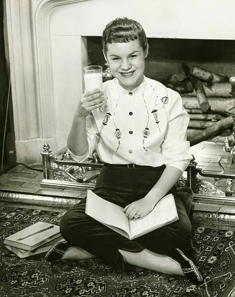 Young woman sitting by fireplace, holding glass of milk in raised hand, (B&W), portrait