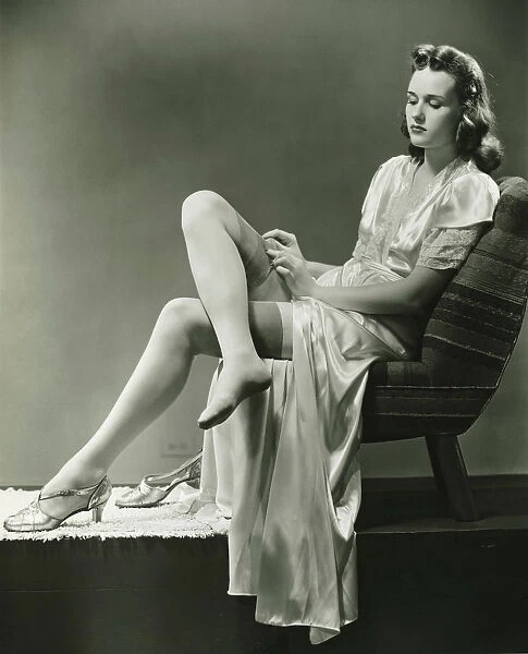 Young woman straightening stockings, sitting on chair in studio, (B&W)