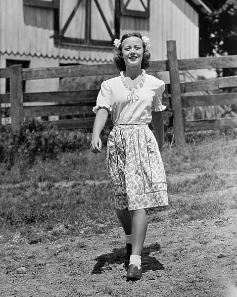 Young woman walking on dirt road by farmhouse, (B&W)
