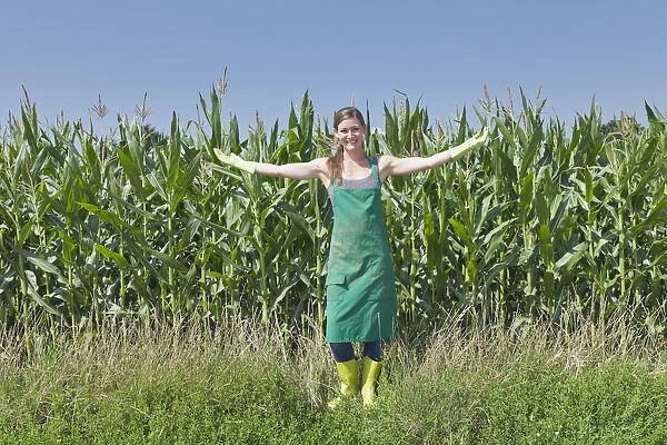 Young woman in work clothes standing with outstretched arms in front of a maize field, Baden-Wurttemberg, Germany