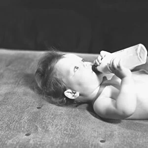 Baby (6-9 months) lying on bed, drinking from bottle, (B&W)