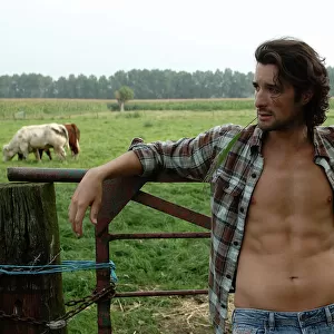 Bare-chested cowboy leaning on a cow gate