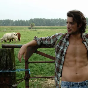 Bare-chested cowboy leaning on a cow gate