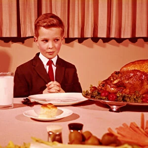 Boy sitting at table with hands folded for grace prayer, looking at roast turkey
