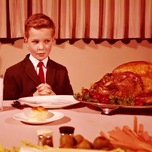 Boy sitting at table with hands folded for grace prayer, looking at roast turkey