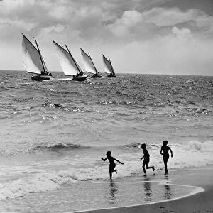 Three boys running along beach, following four sailboats out on ocean