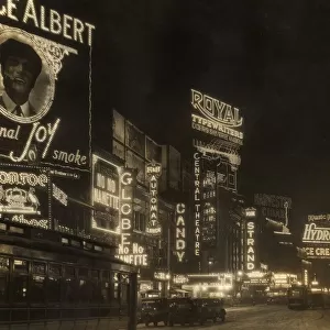 circa 1926: Automobiles and a streetcar pass illuminated theater marquees and billboards