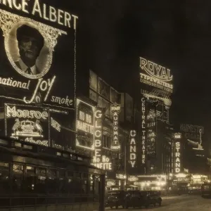 circa 1926: Automobiles and a streetcar pass illuminated theater marquees and billboards