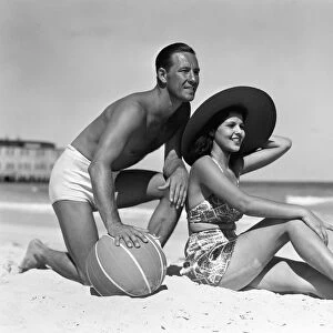 Couple on beach, man leaning on ball, woman sitting look out to ocean, wearing sun hat